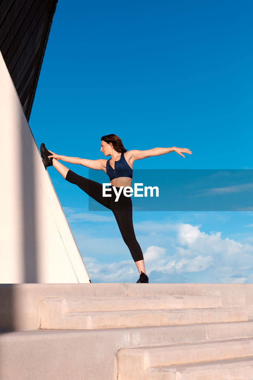 Full body of sporty female in activewear split and stretching leg on wall during outdoor training against blue sky on summer day