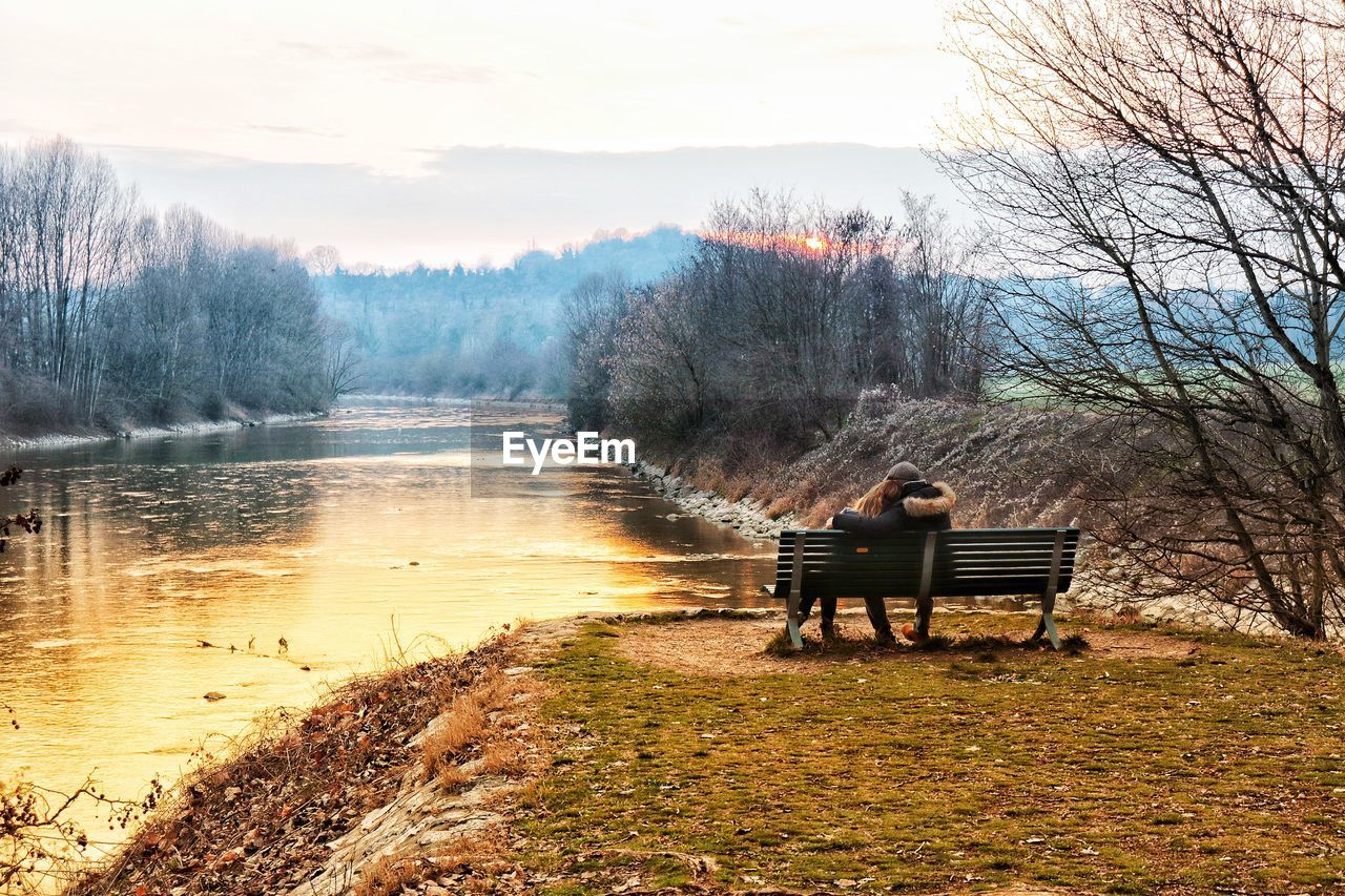 Rear view of couple sitting on bench at lakeshore against sky during sunset