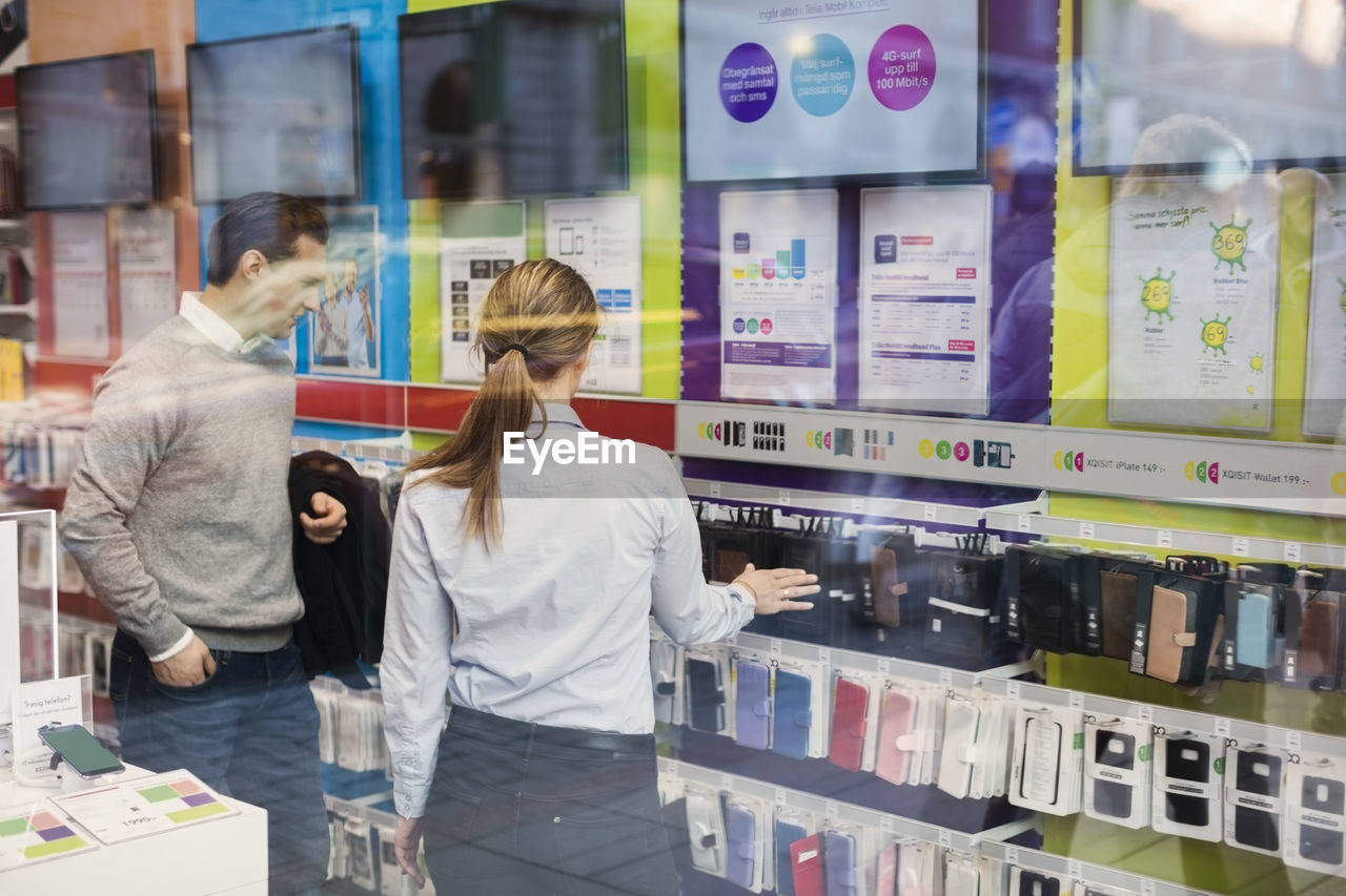 Saleswoman helping customer in buying phone cover seen through glass