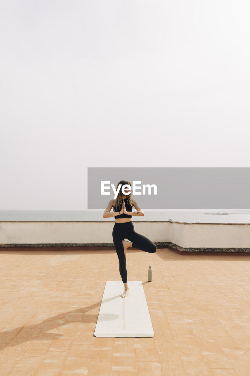 Young woman in sportswear doing tree pose on rooftop against sky