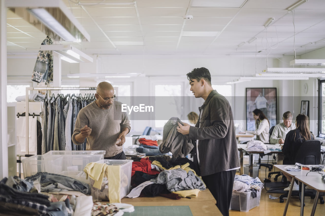 Male fashion designers discussing with each other while sorting recycled clothes at workshop