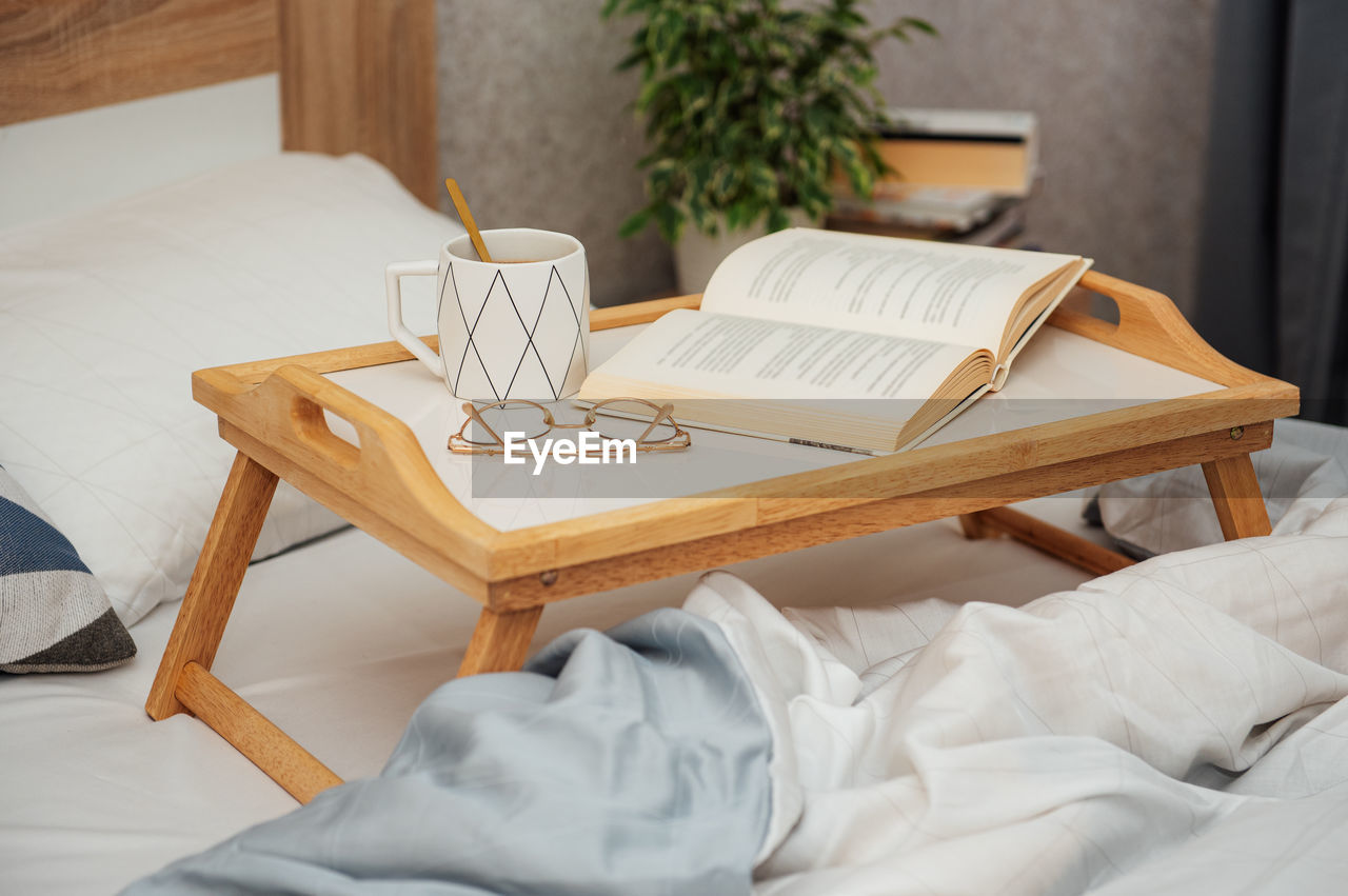 Cup of coffee, book and glasses on bed with soft blanket indoors