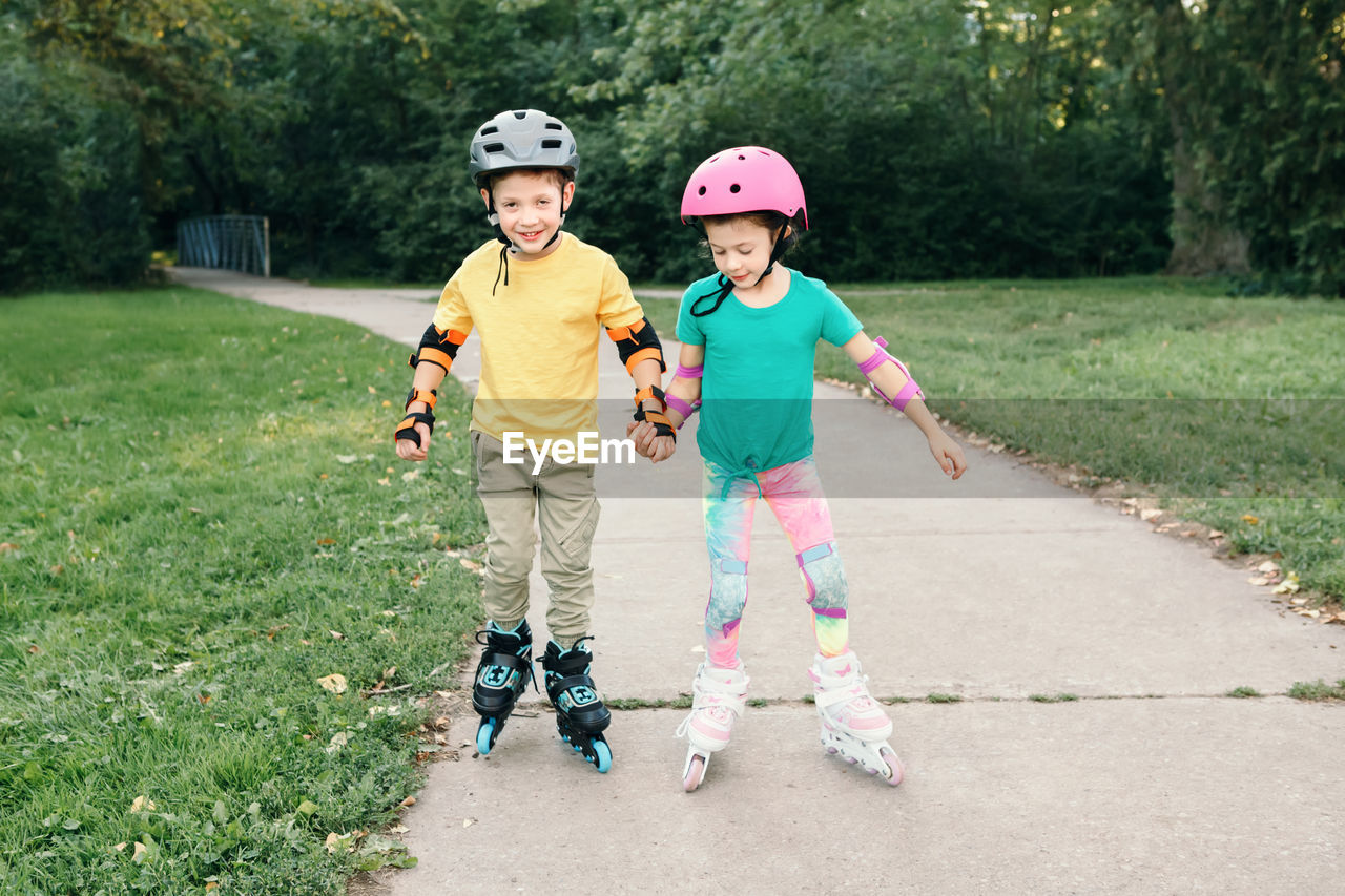 Happy caucasian friends boy and girl in helmets riding on roller skates in park on summer day. 