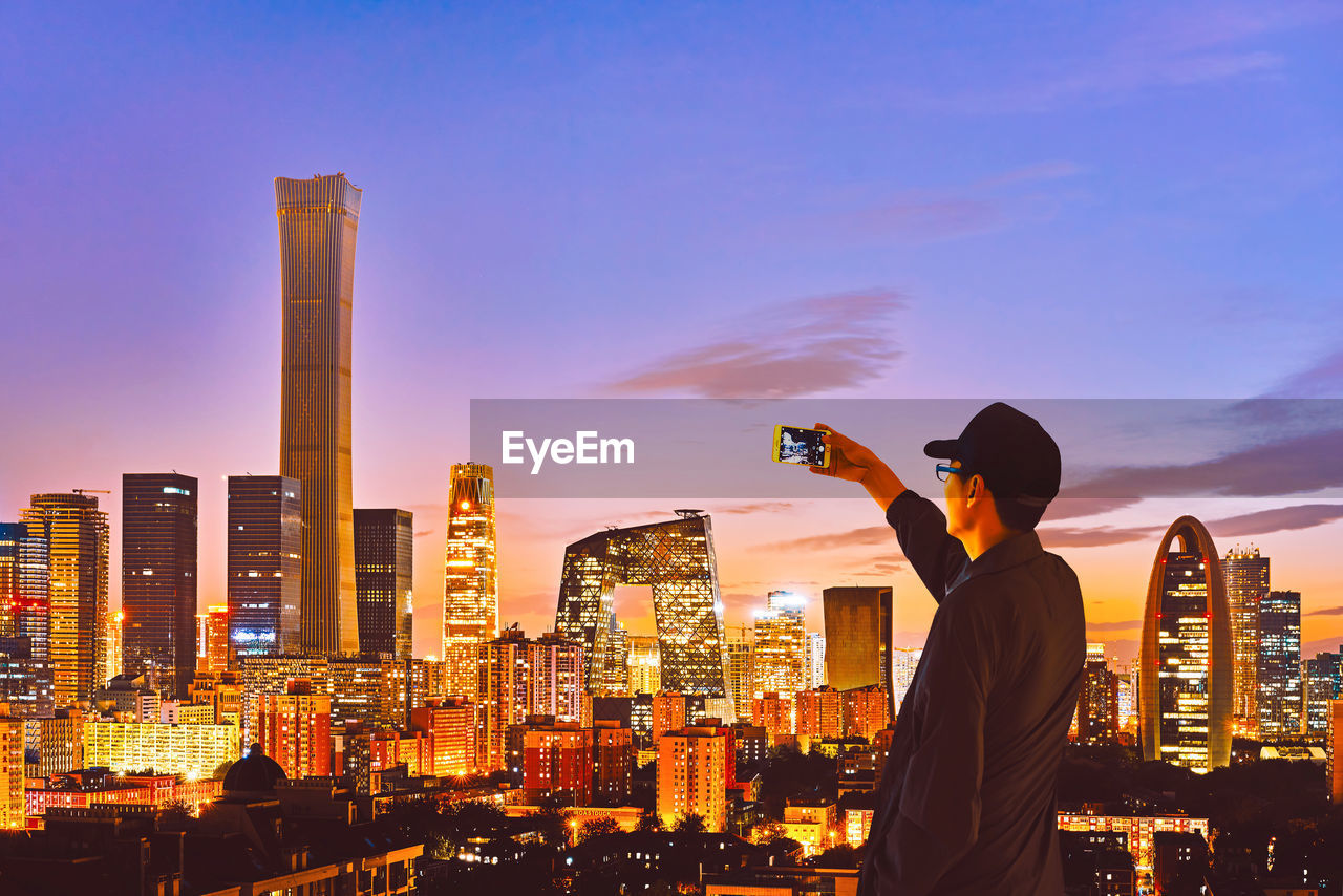 Rear view of man photographing cityscape against sky during sunset