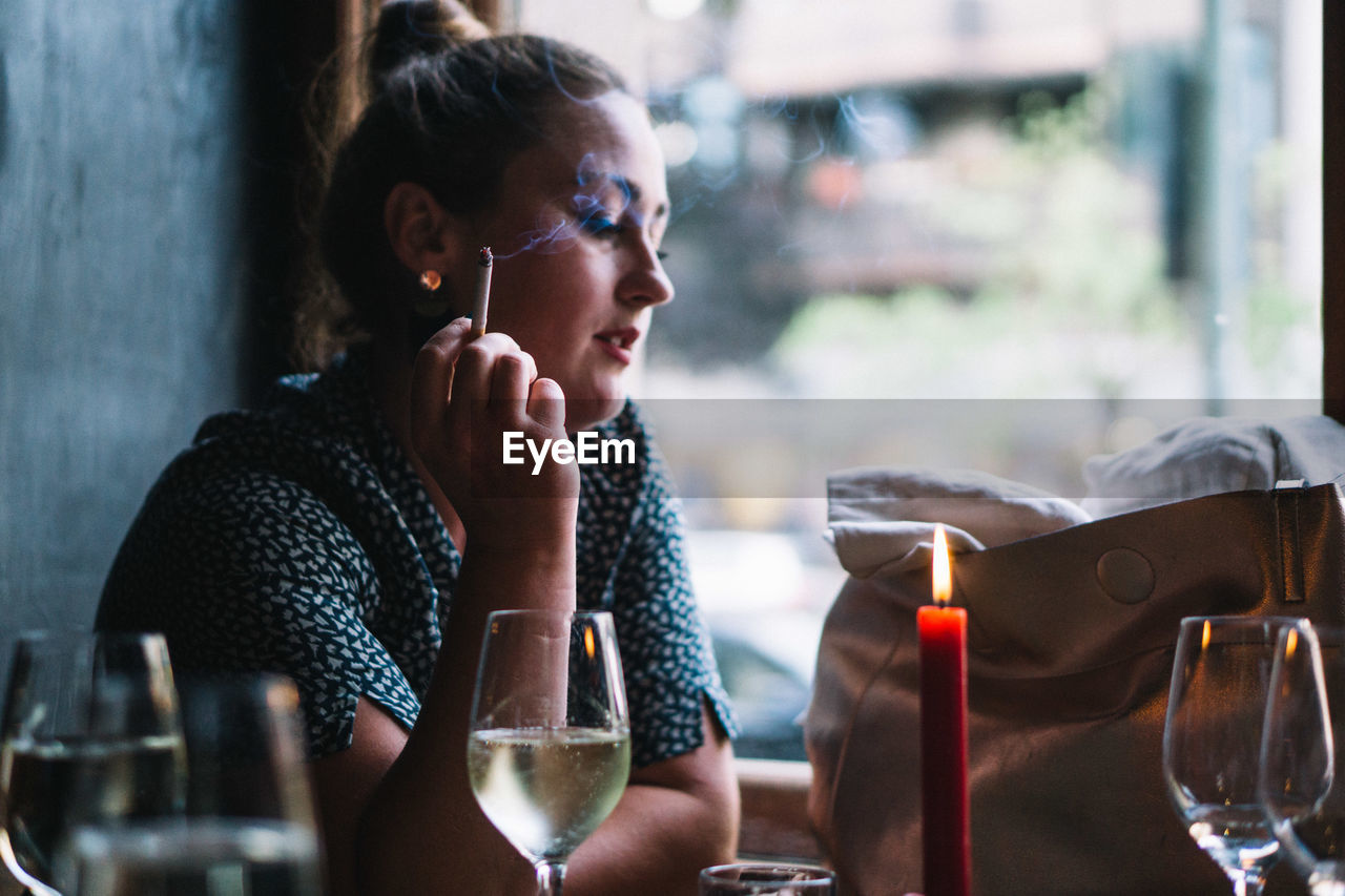 Side view of woman smoking cigarette while sitting with wineglass at restaurant table