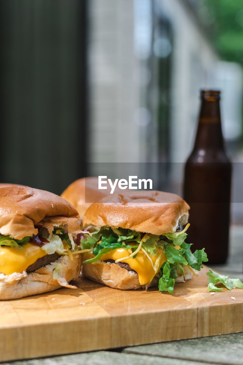 A set of cheeseburgers on a wooden plate and a couple of beers on an outdoor table. suburban.