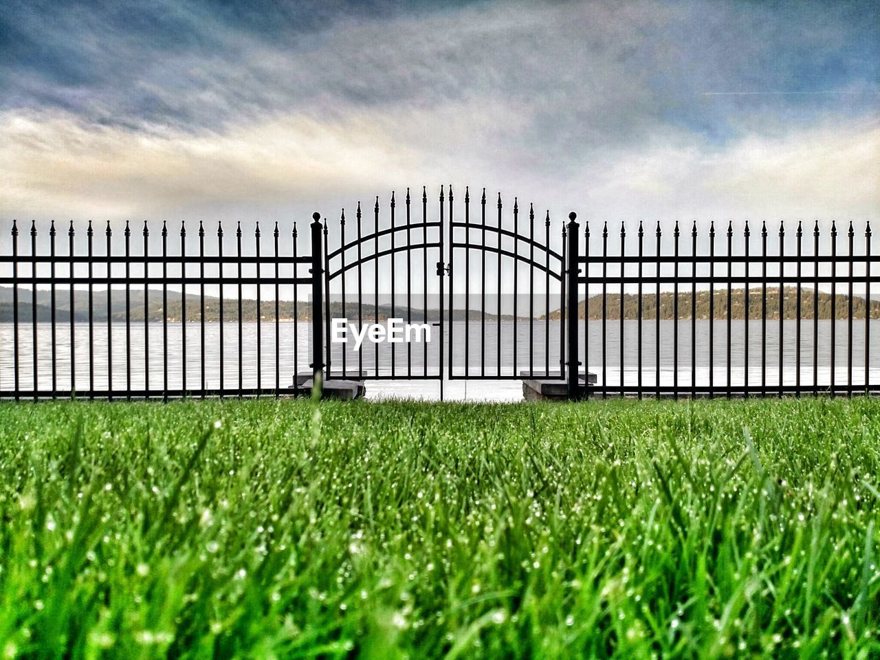 Metallic gate on grassy field by lake against cloudy sky