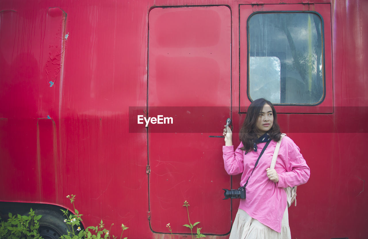 Woman looking away while standing against red vehicle