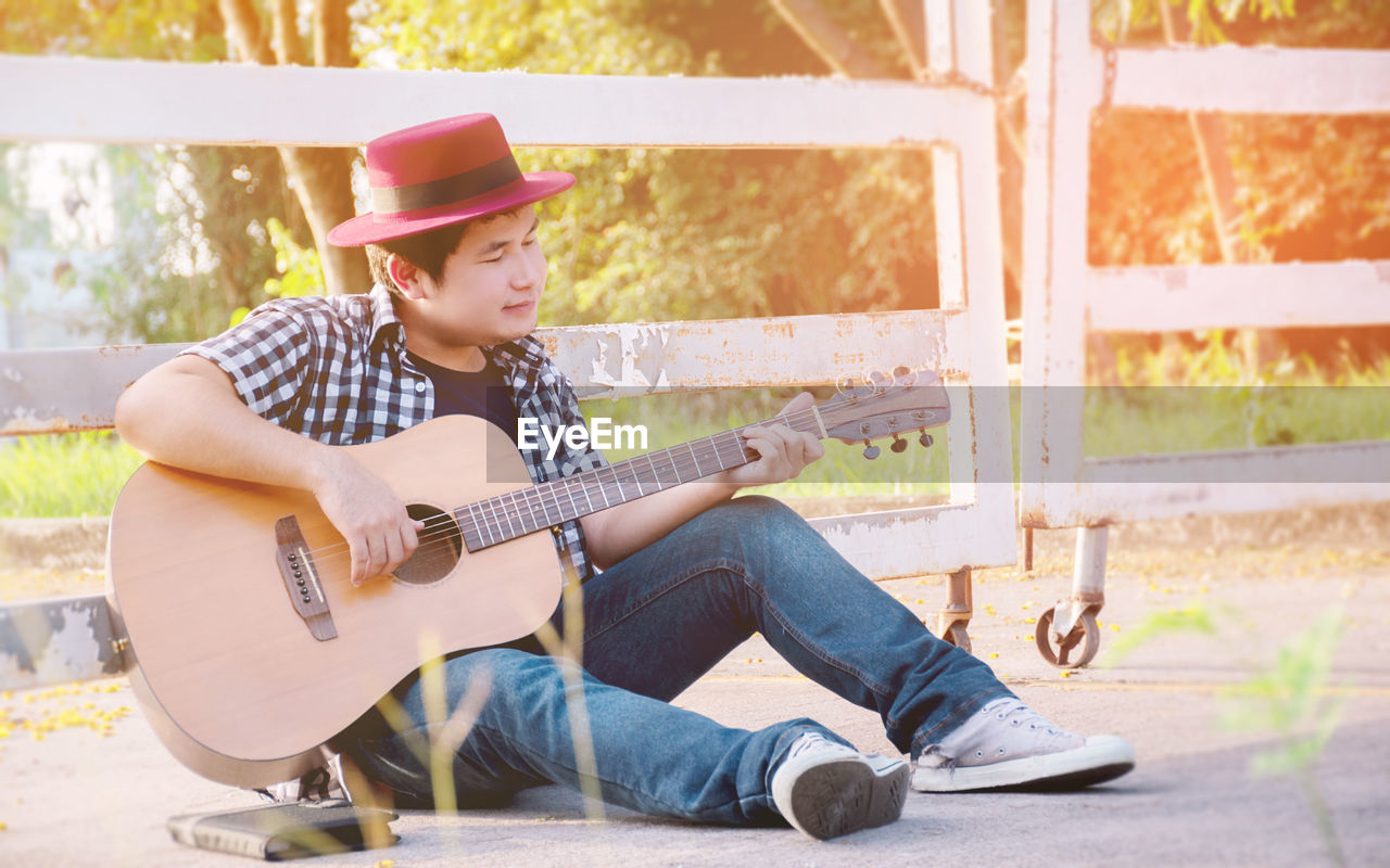 Full length of man playing guitar while sitting against fence