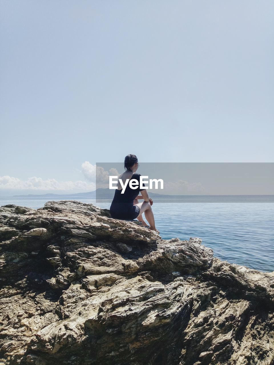 Young woman sitting on rock by sea against clear sky