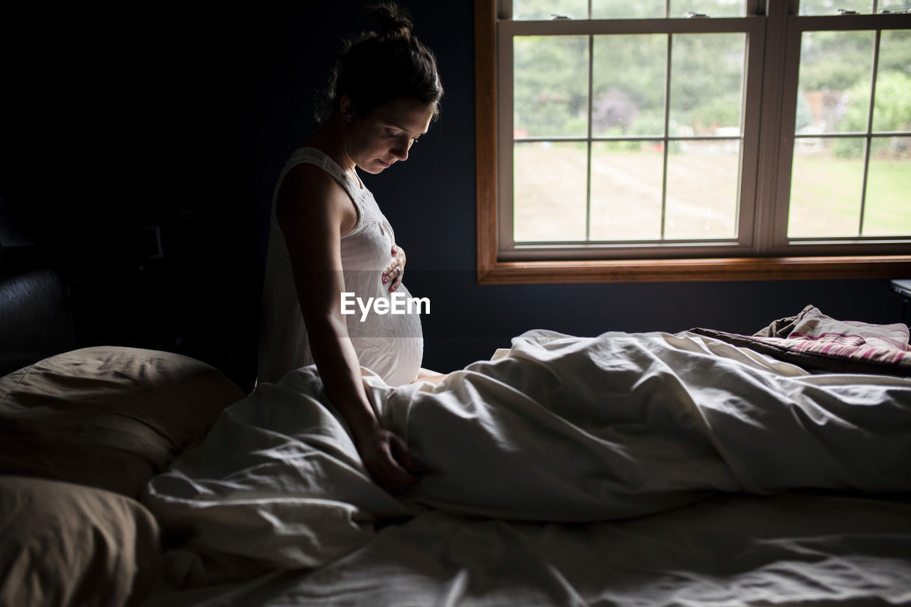 Side view of pregnant woman touching belly on bed at home