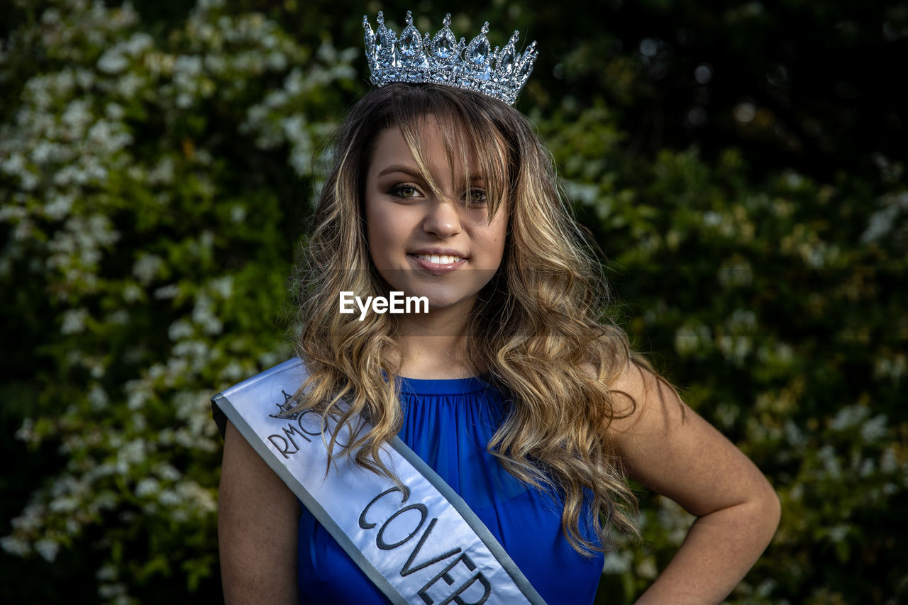 Portrait of smiling young woman wearing crown standing against plants