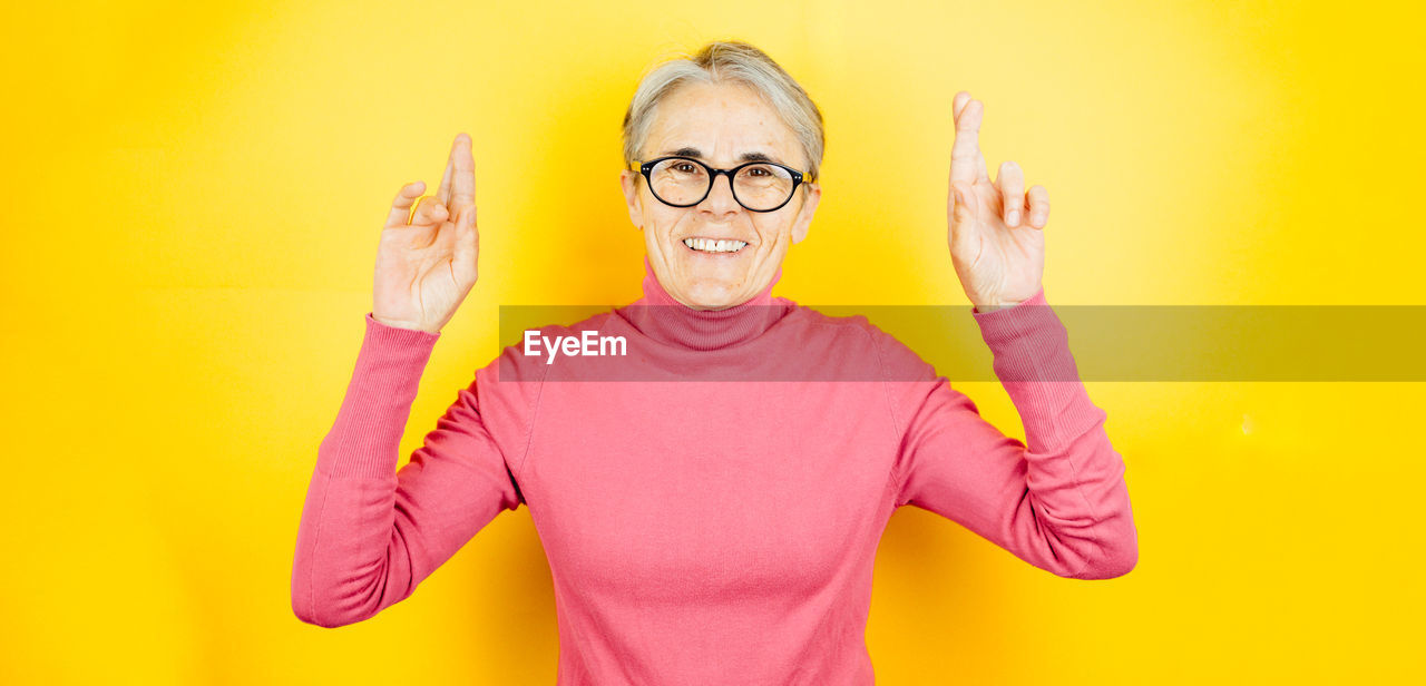 yellow, one person, eyeglasses, glasses, yellow background, portrait, adult, studio shot, colored background, finger, pink, waist up, humor, indoors, front view, fun, emotion, looking at camera, smiling, arm, happiness, casual clothing, human face, hand, gesturing, standing, t-shirt, facial expression, person, clothing, sign language, women, copy space, arms raised, individuality, positive emotion