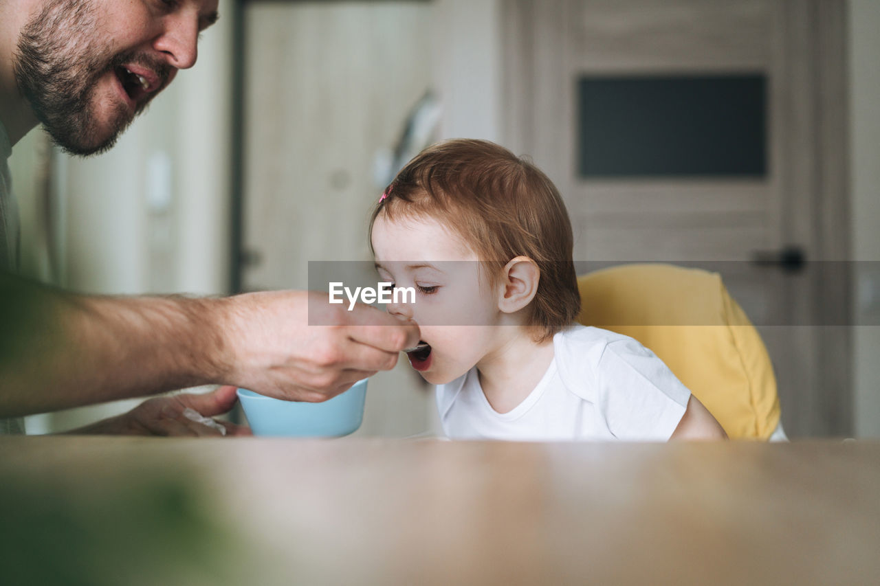 Happy father young man feeds baby girl little daughter in kitchen at home