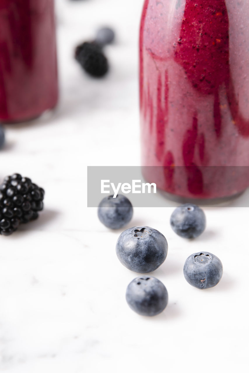 Details of blueberries beside a glass bottle with smoothie over a marble surface.