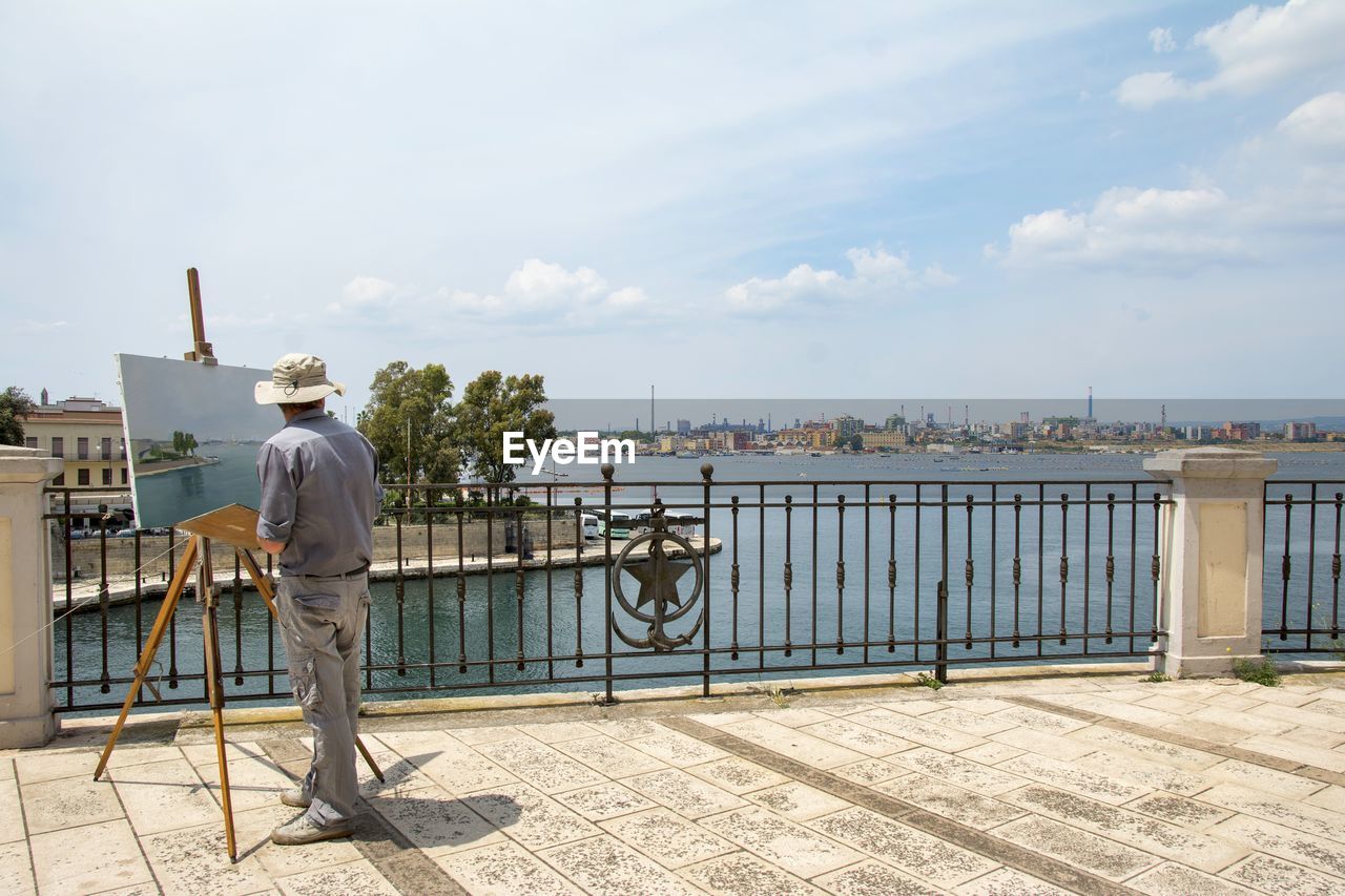 REAR VIEW OF MAN STANDING ON RAILING AGAINST SKY