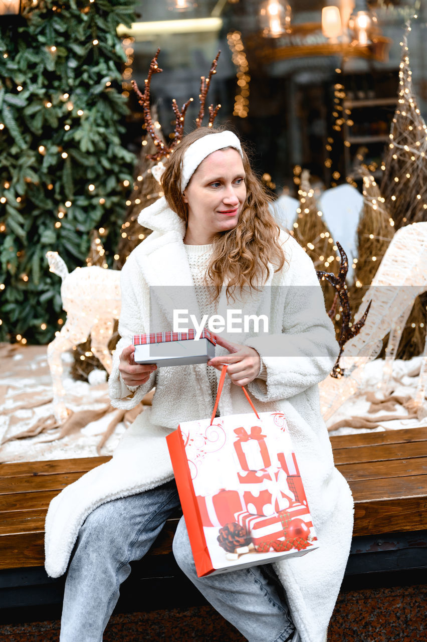 Woman in white fur coat sits on a bench outside. she holds a white mug with a christmas print