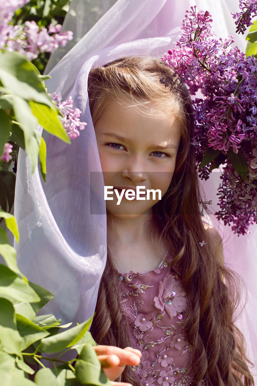 Caucasian girl child seven years old in a purple dress stands in nature with a wreath of lilacs