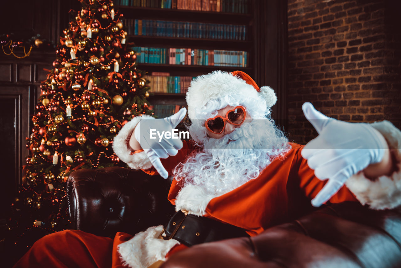 Portrait of man in santa claus costume gesturing while sitting by christmas tree at home