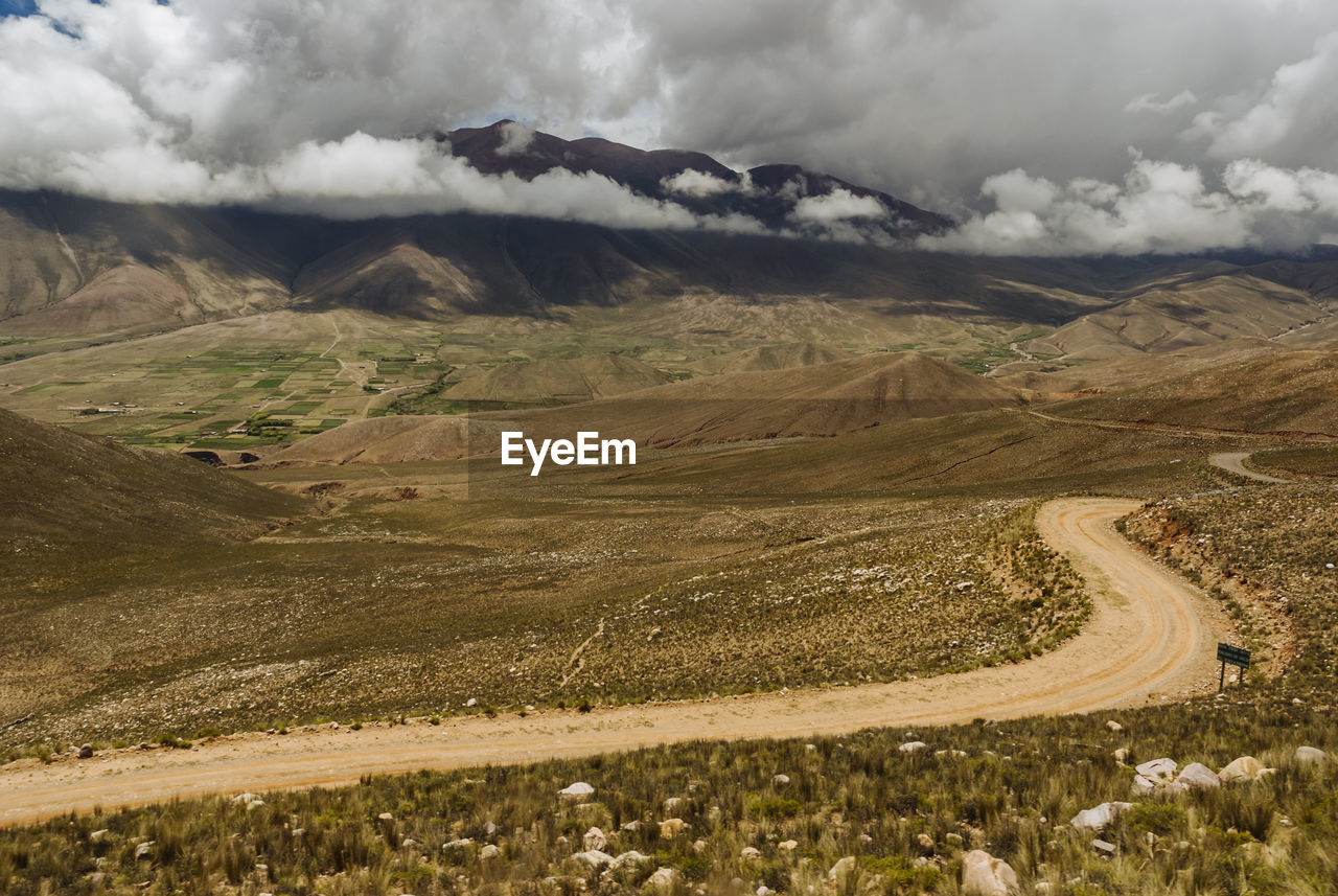 Dirt road on field against cloudy sky