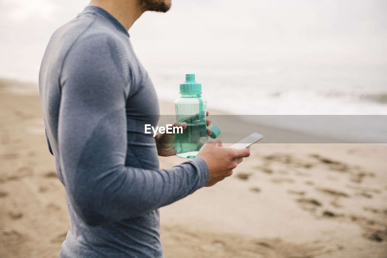 Sportive young man with cell phone and drinking bottle on the beach