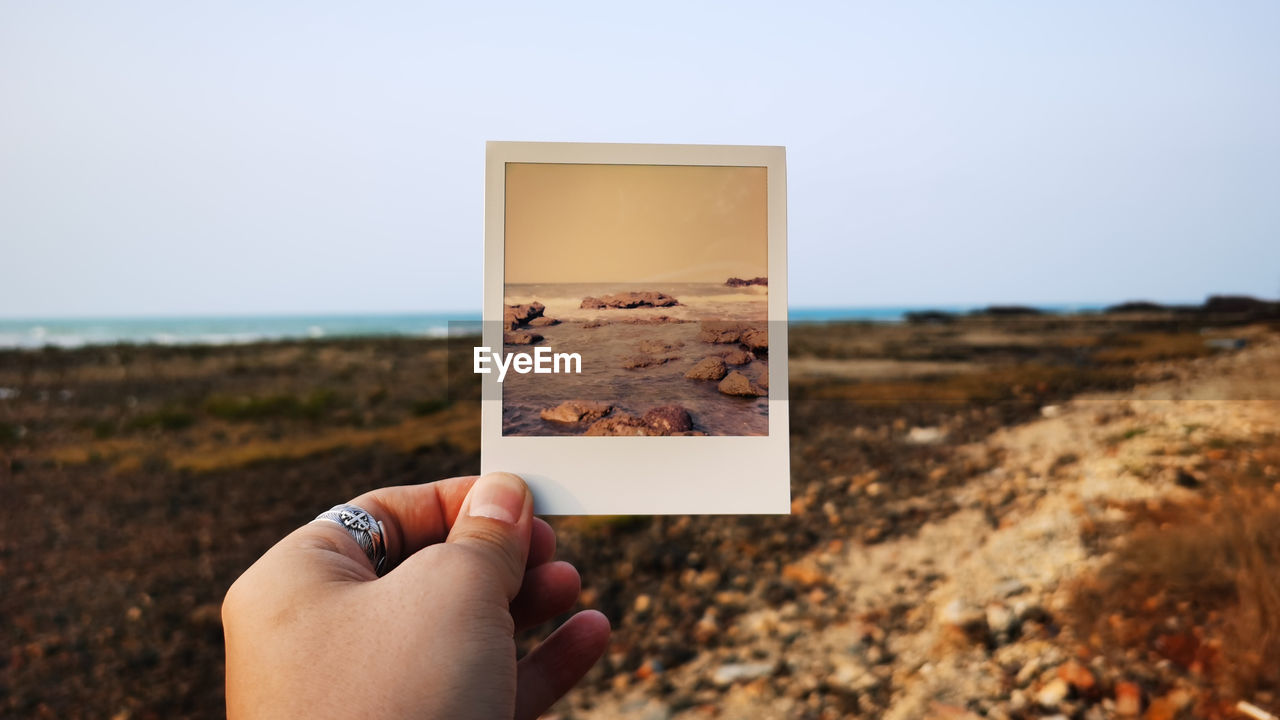 Cropped image of person holding instant print transfer on beach at sea against sky