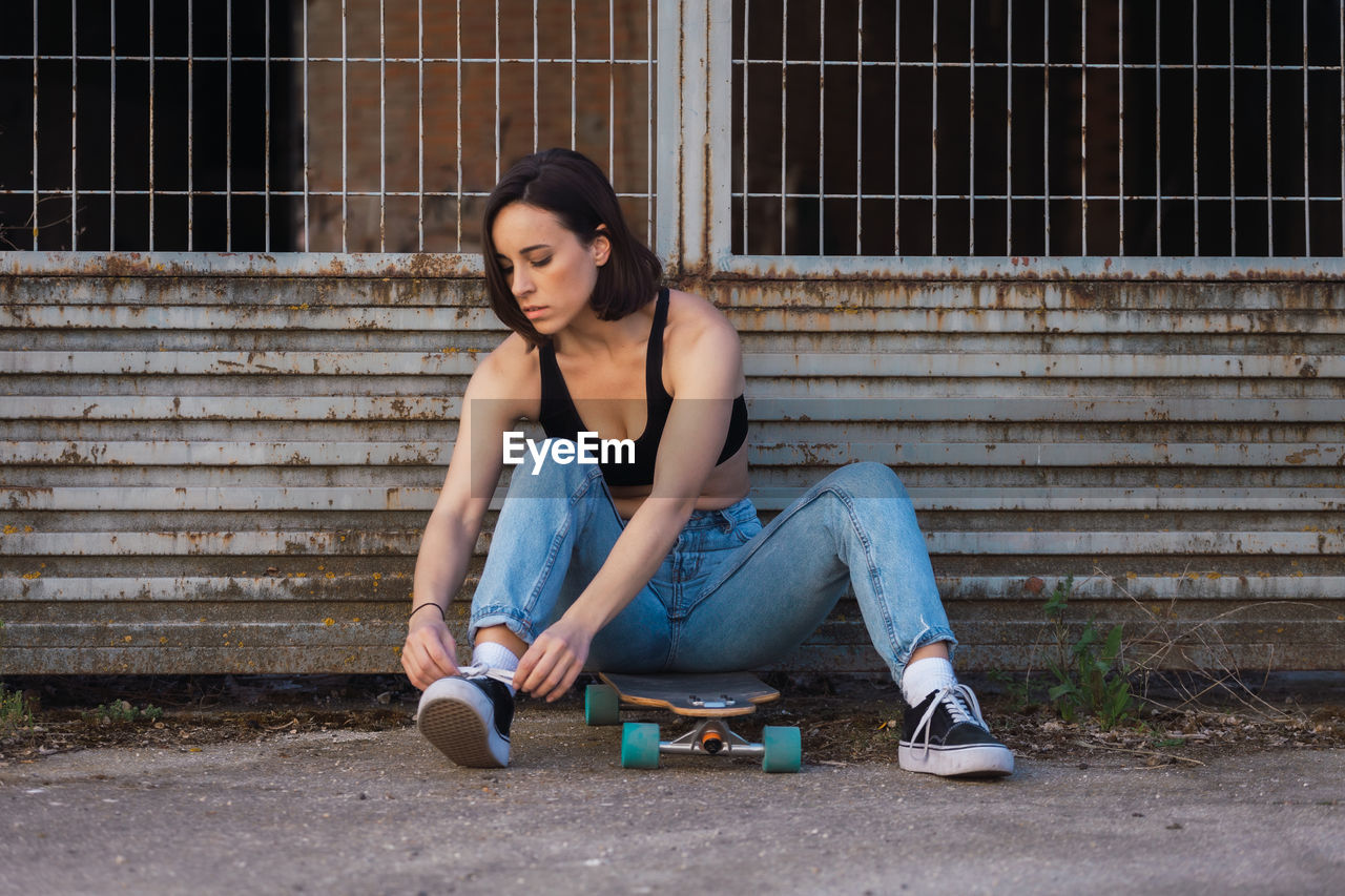 Full length of young woman sitting on skateboard tying shoelace outdoors