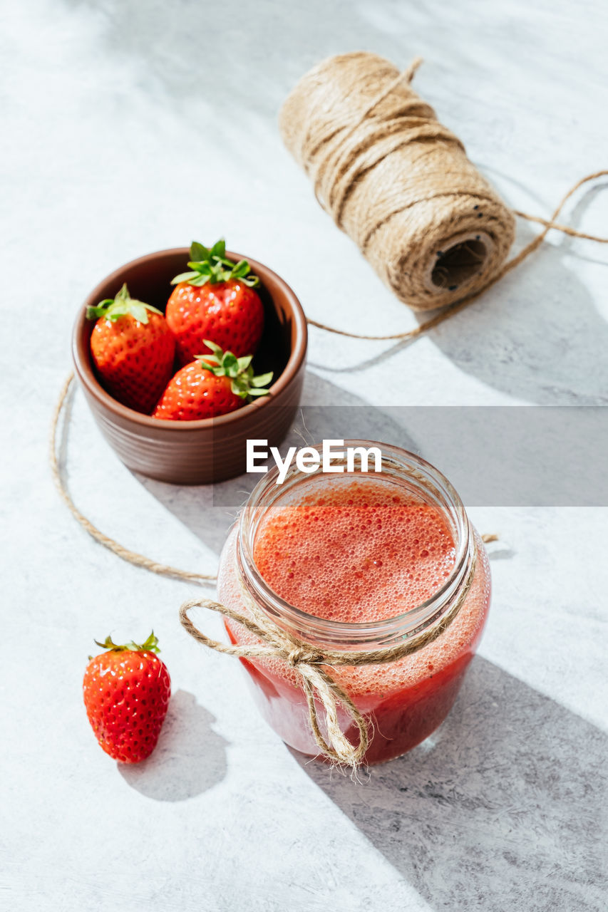 From above composition with fresh homemade strawberry juice in glass jar wrapped with twine placed on marble surface with whole berries