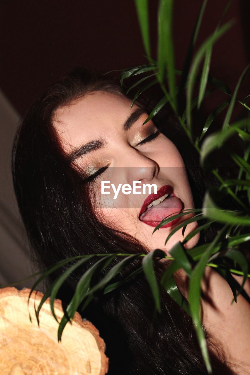 Close-up of young woman with eyes closed sticking out tongue by plants