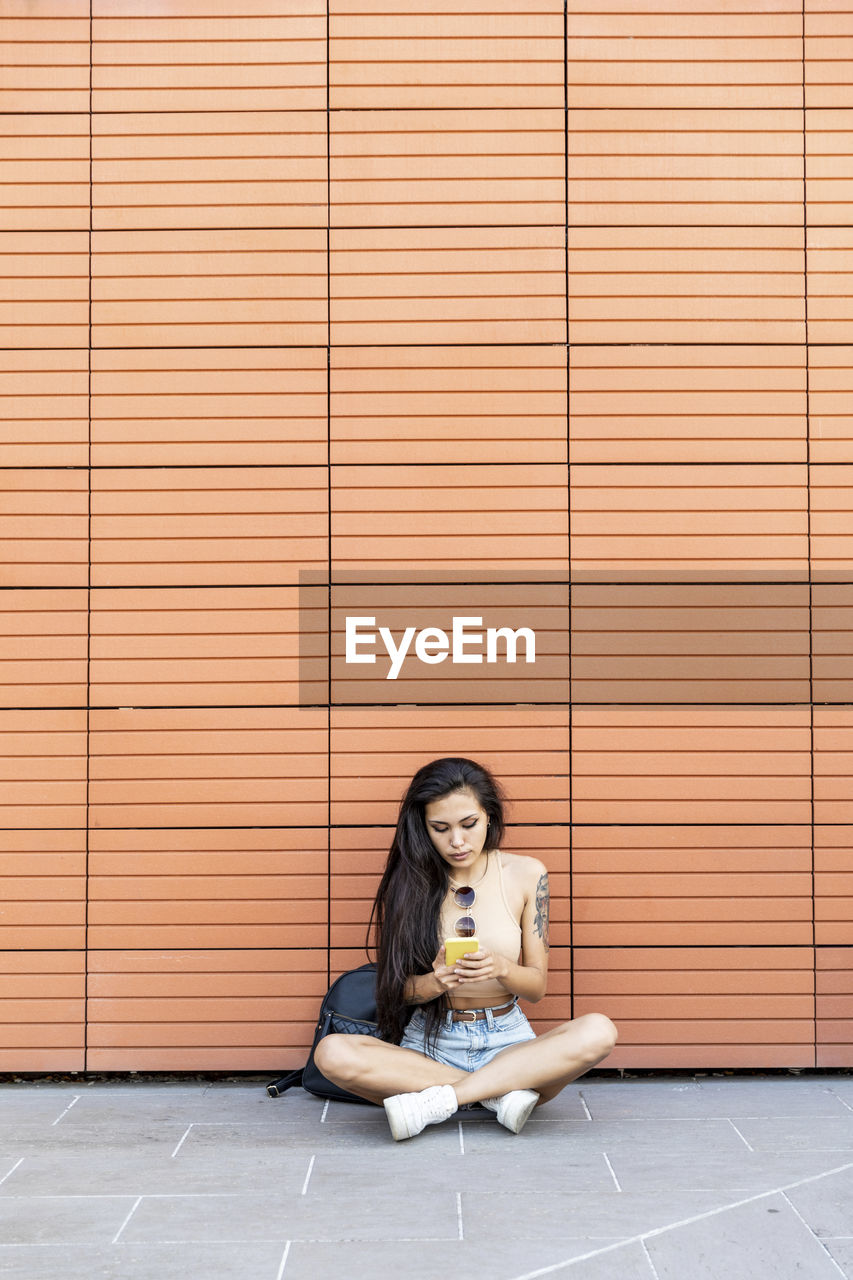 Young woman using mobile phone while sitting against wall