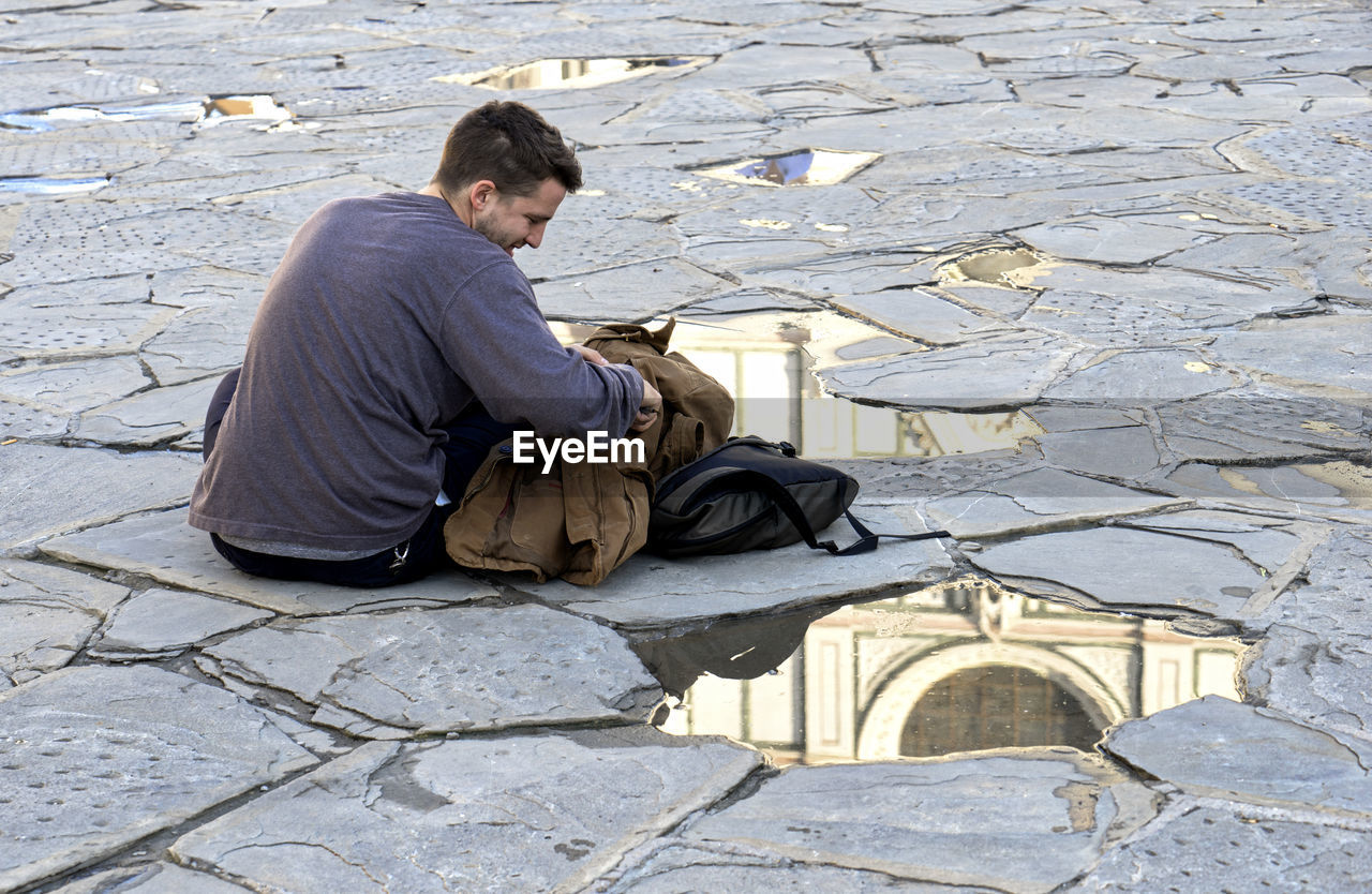 MAN SITTING ON STREET AGAINST STONE WALL