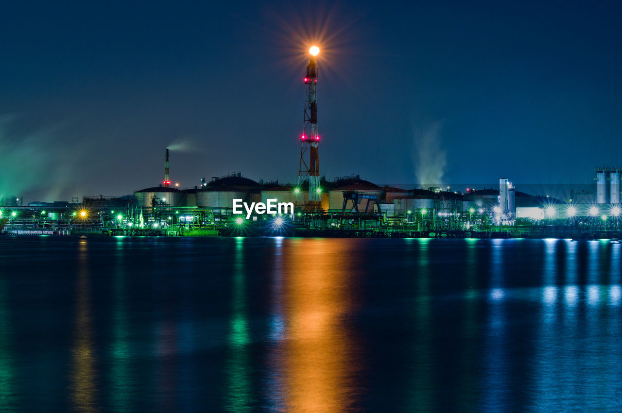 Illuminated factory buildings by sea against sky at night