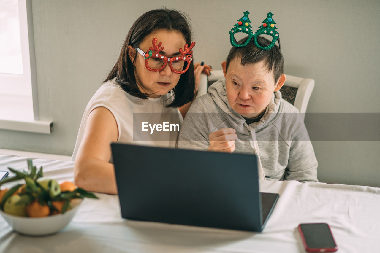 Girl helps elderly woman with down syndrome in funny christmas glasses, make video call from laptop