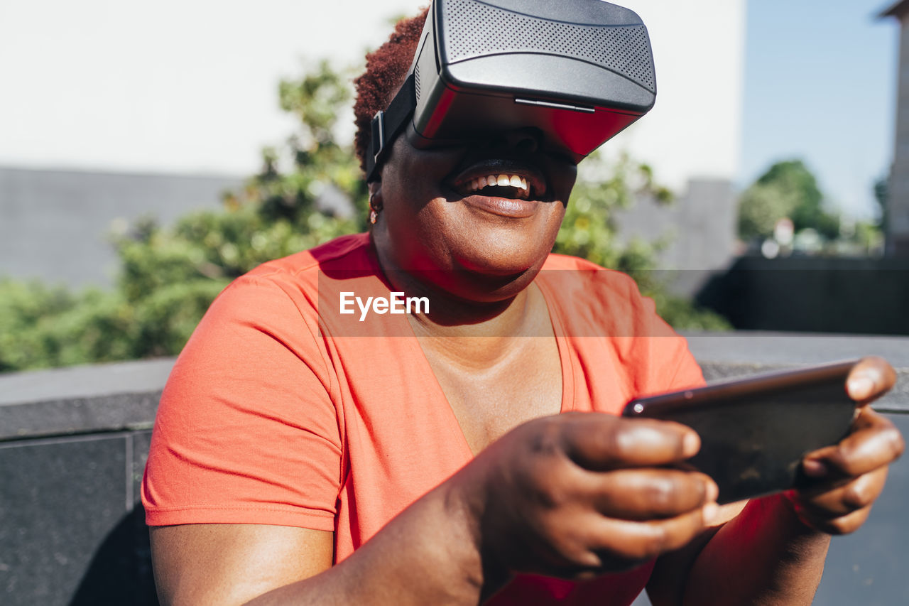 Playful woman wearing virtual reality holding smart phone on sunny day