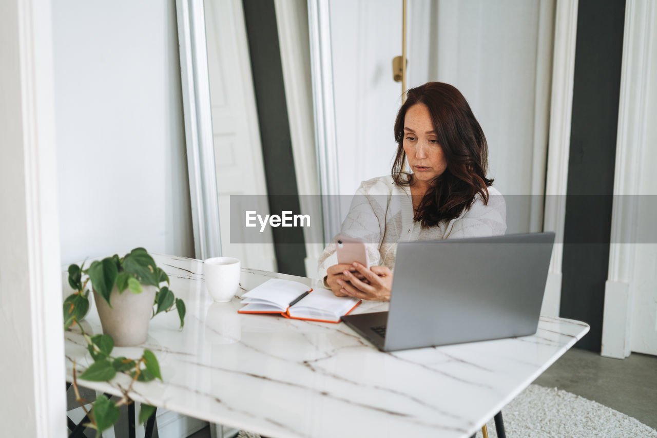 Thinking woman with long hair in white cardigan working on laptop using mobile phone in office