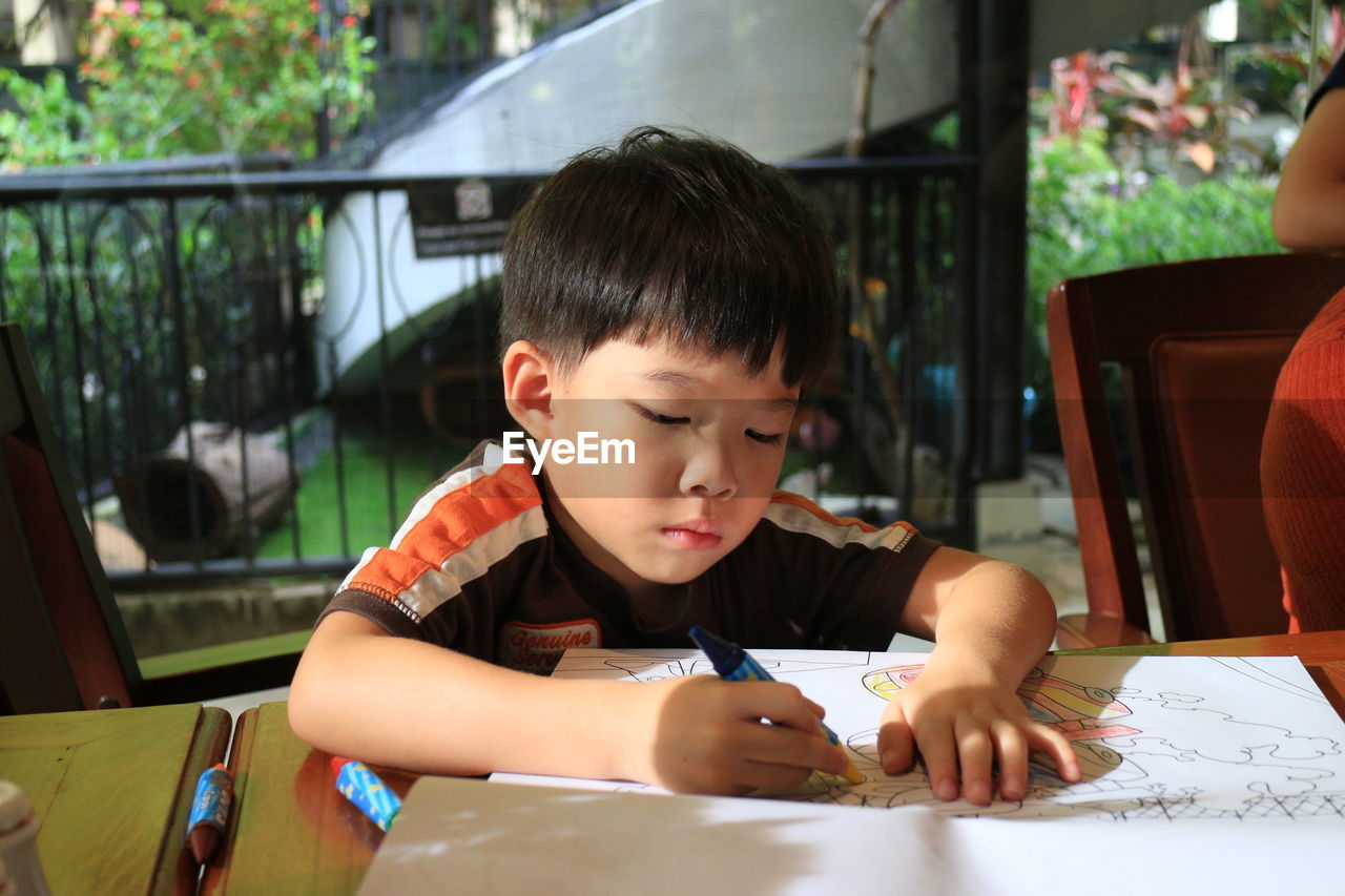 Boy drawing on book while sitting at table in porch