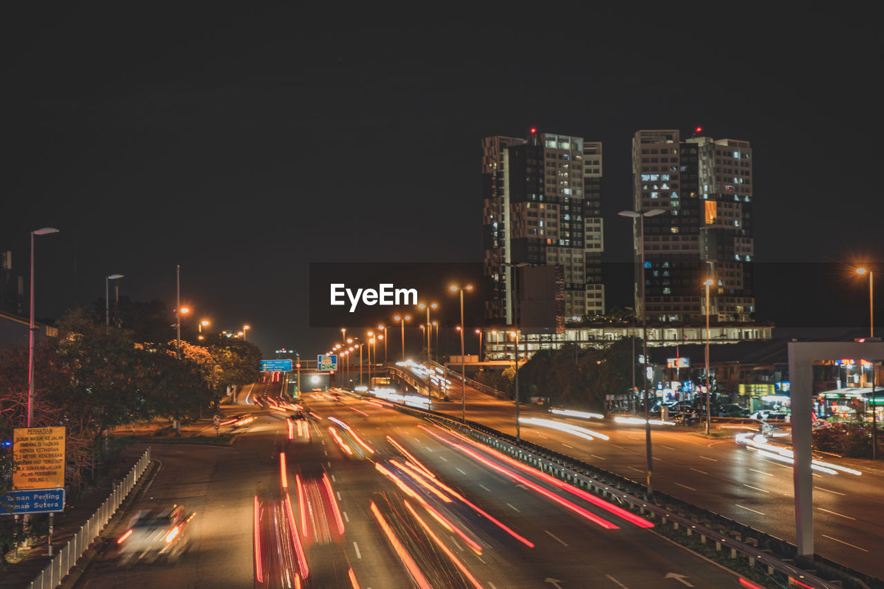 Light trails on road amidst buildings against sky at night