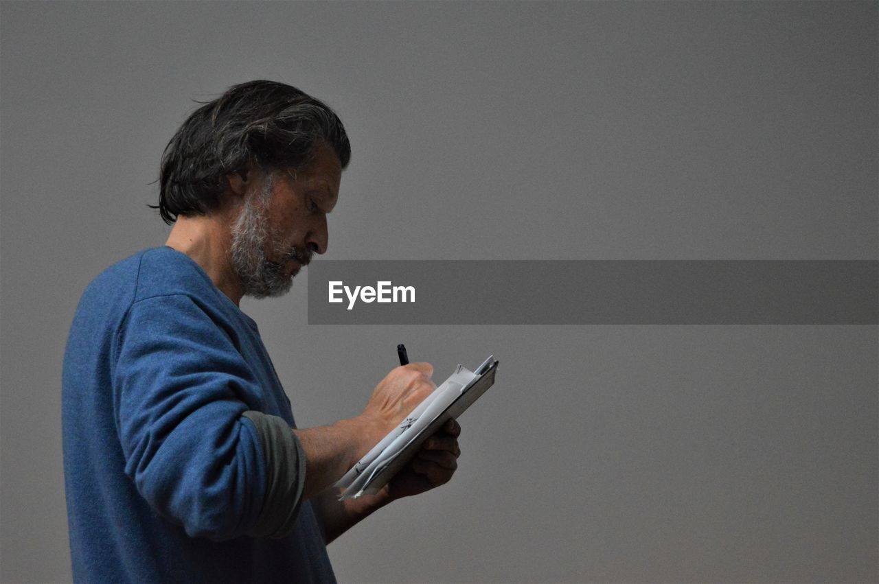 Side view of mature man writing while standing against gray background