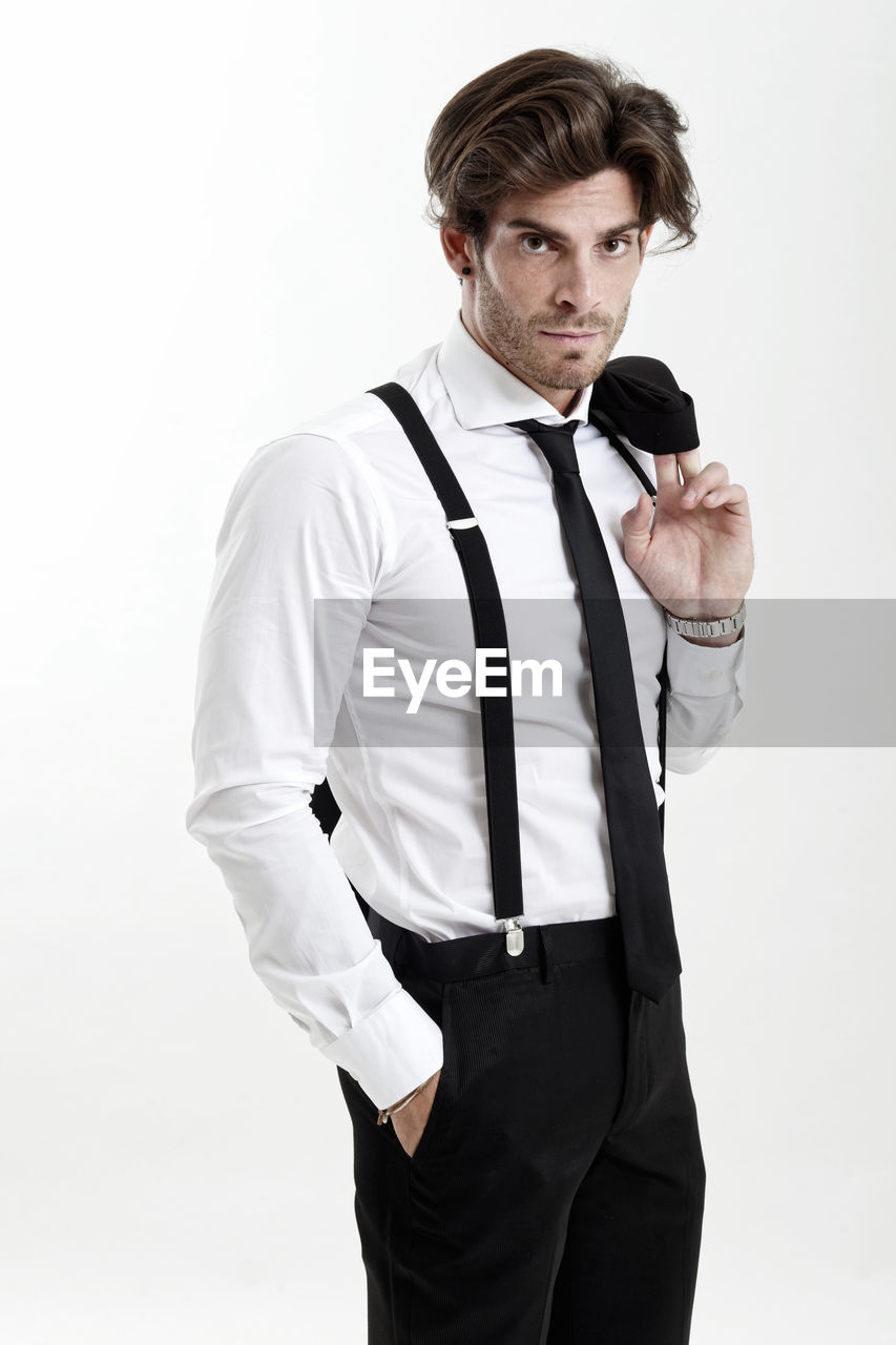 Portrait of man wearing suspenders standing with hands in pockets against white background