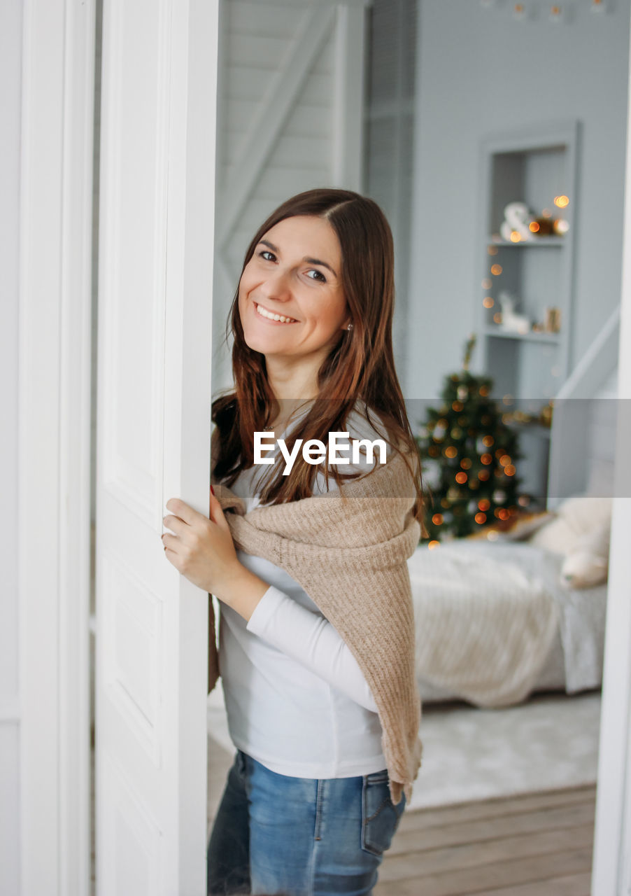 PORTRAIT OF SMILING YOUNG WOMAN STANDING AGAINST WALL AT HOME