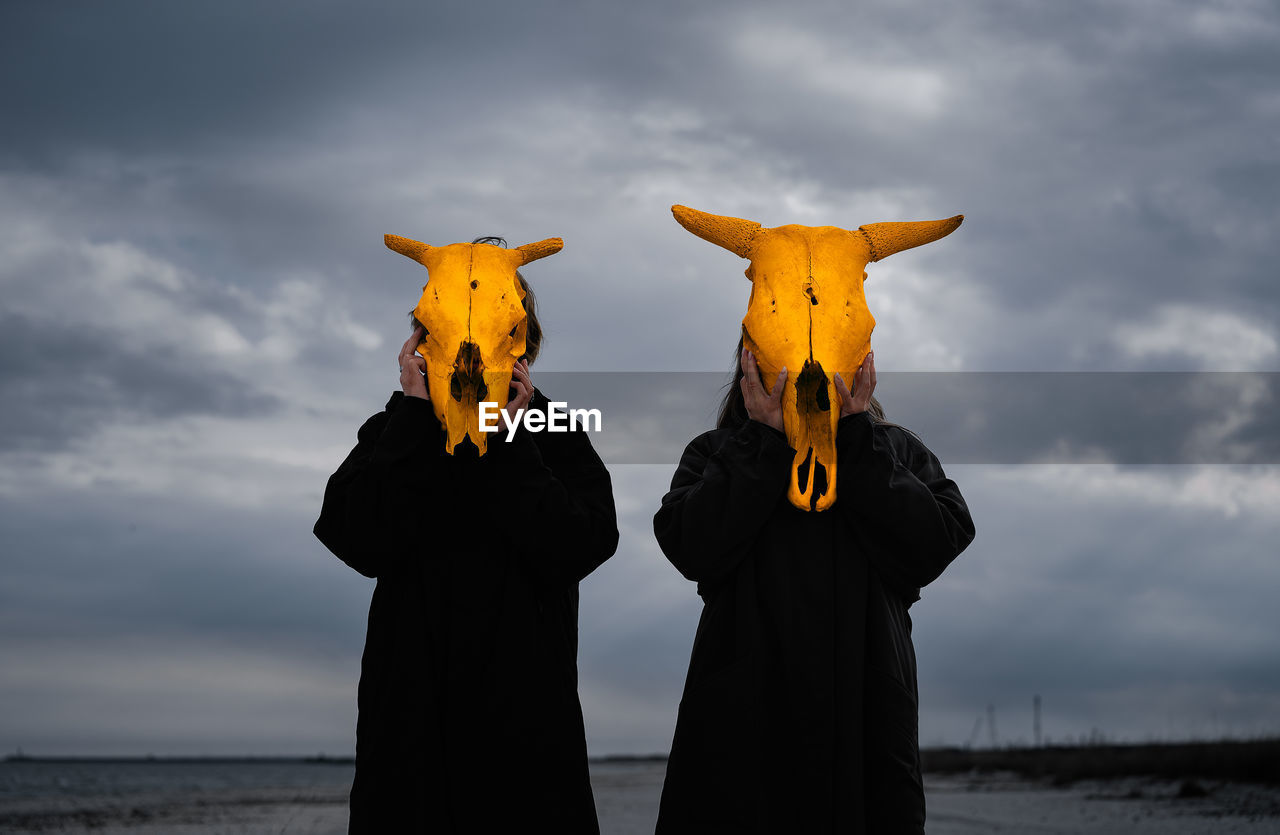 People with yellow mask standing by sea against cloudy sky