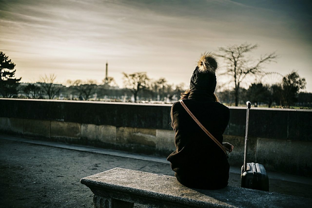 Rear view of woman sitting on bench with suitcase against sky