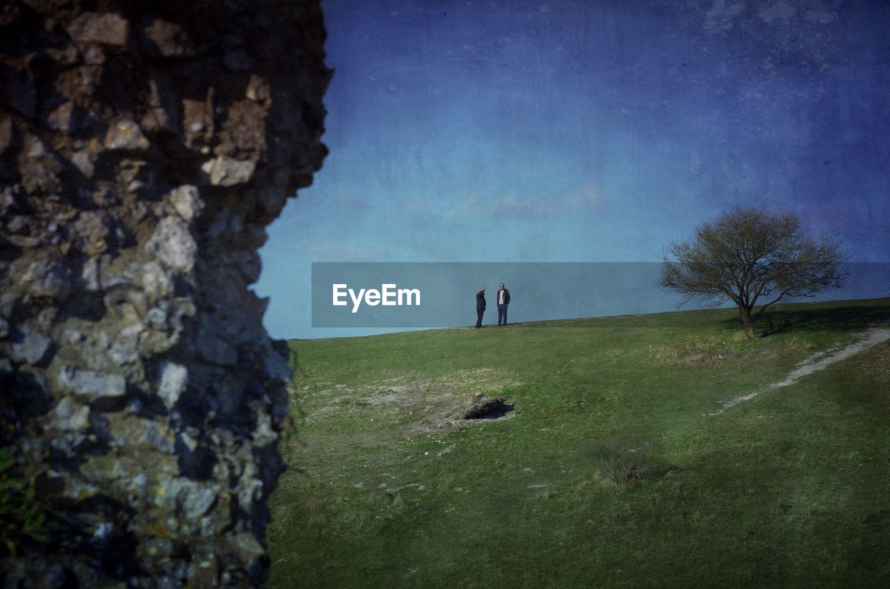 Distant view of men standing on grassy hill against blue sky