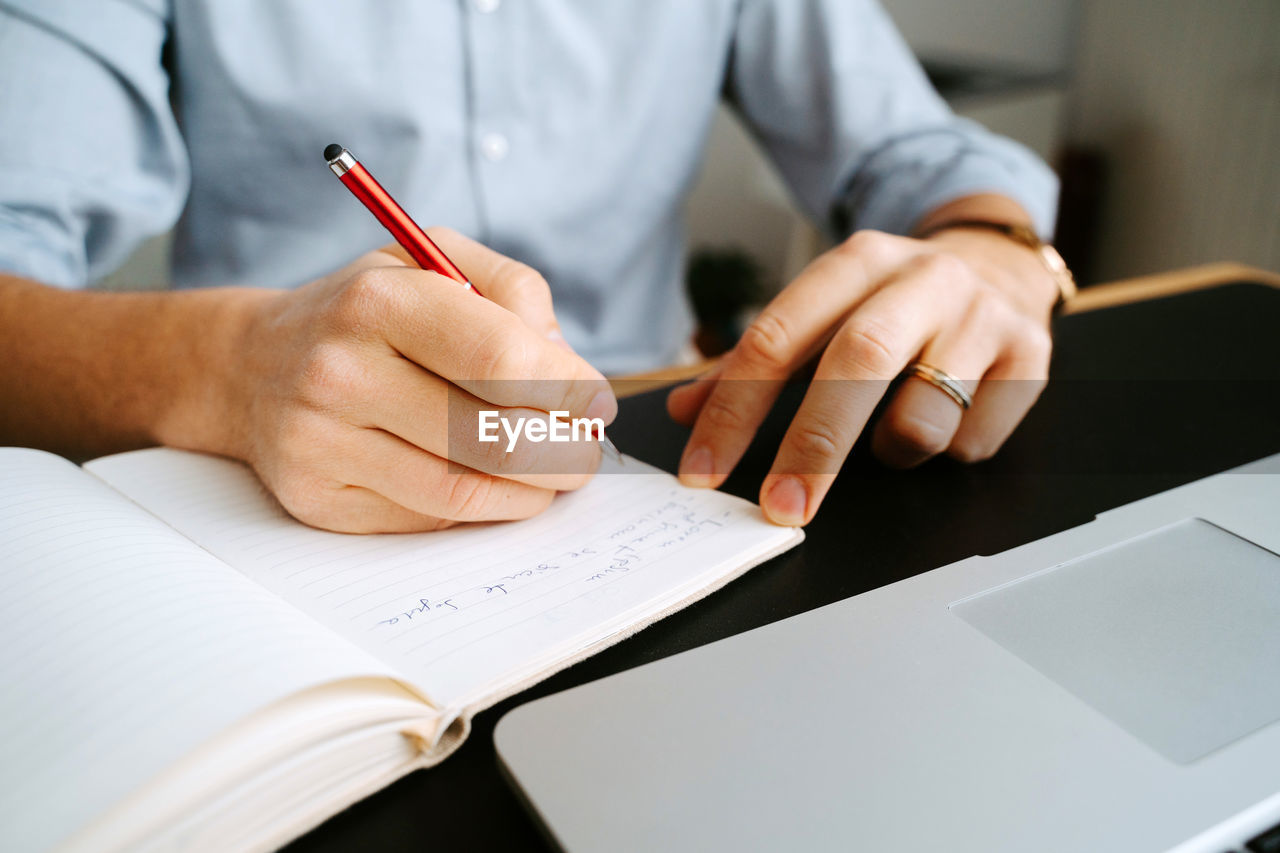 Unrecognizable crop male entrepreneur sitting at table and writing in notebook while planning business project