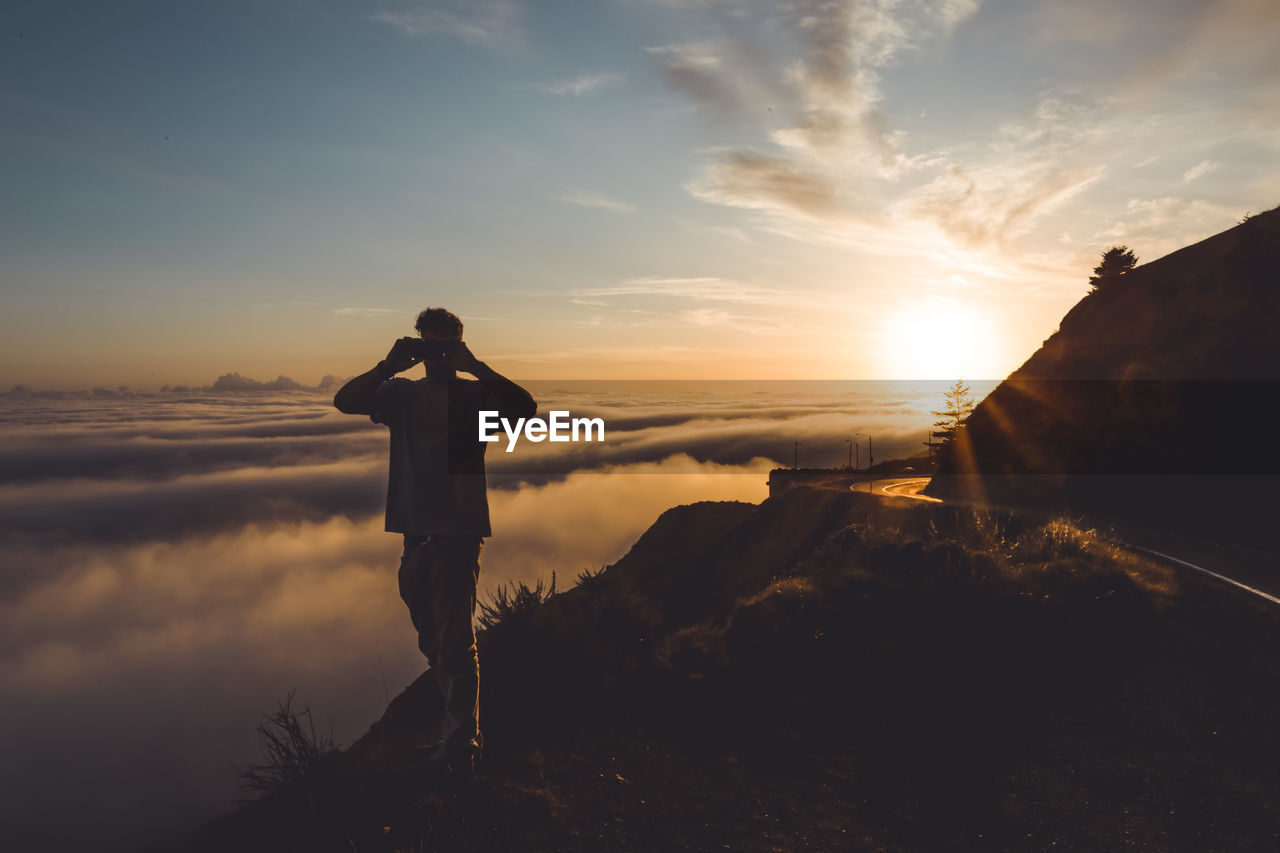 Young male photographing sunset above clouds next to highway