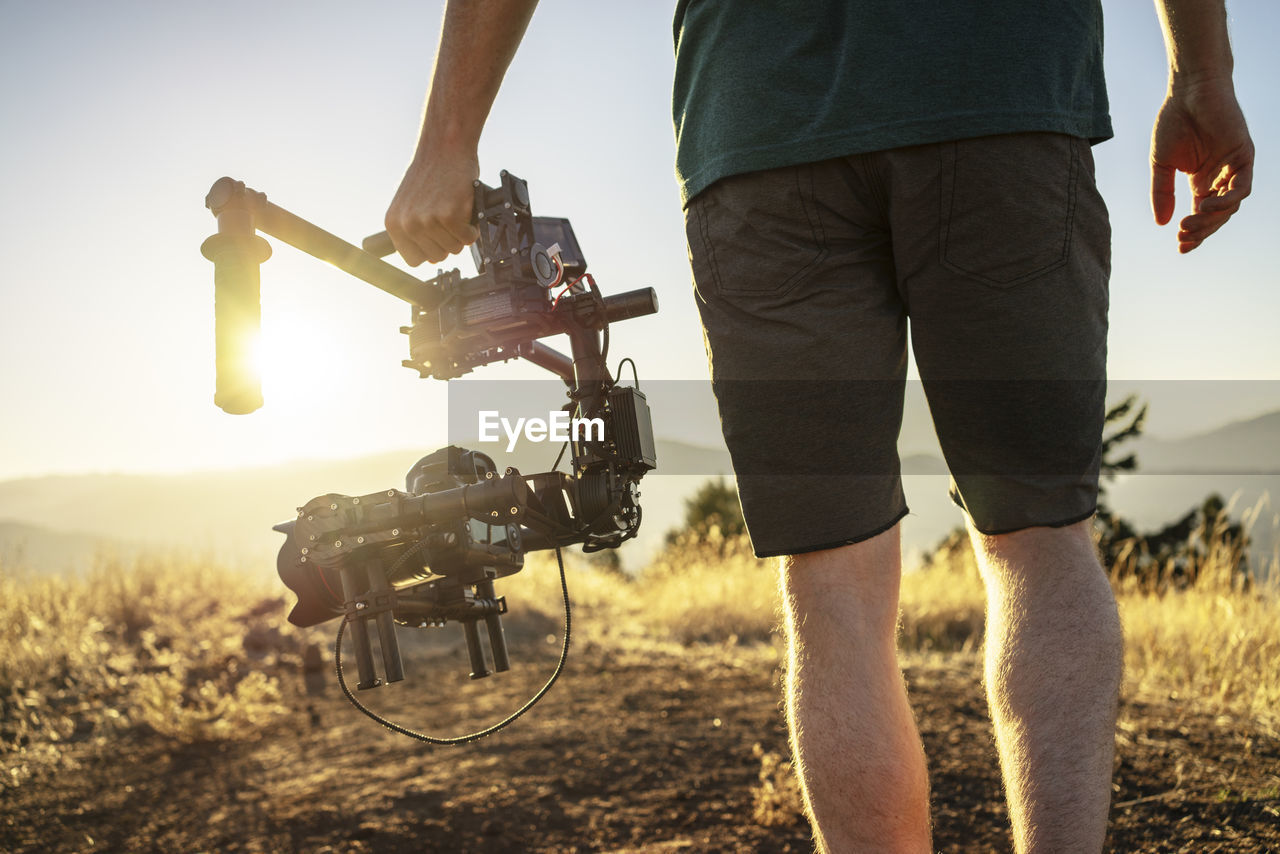 Midsection of man holding shoulder stand camera while standing on mountain during sunny day