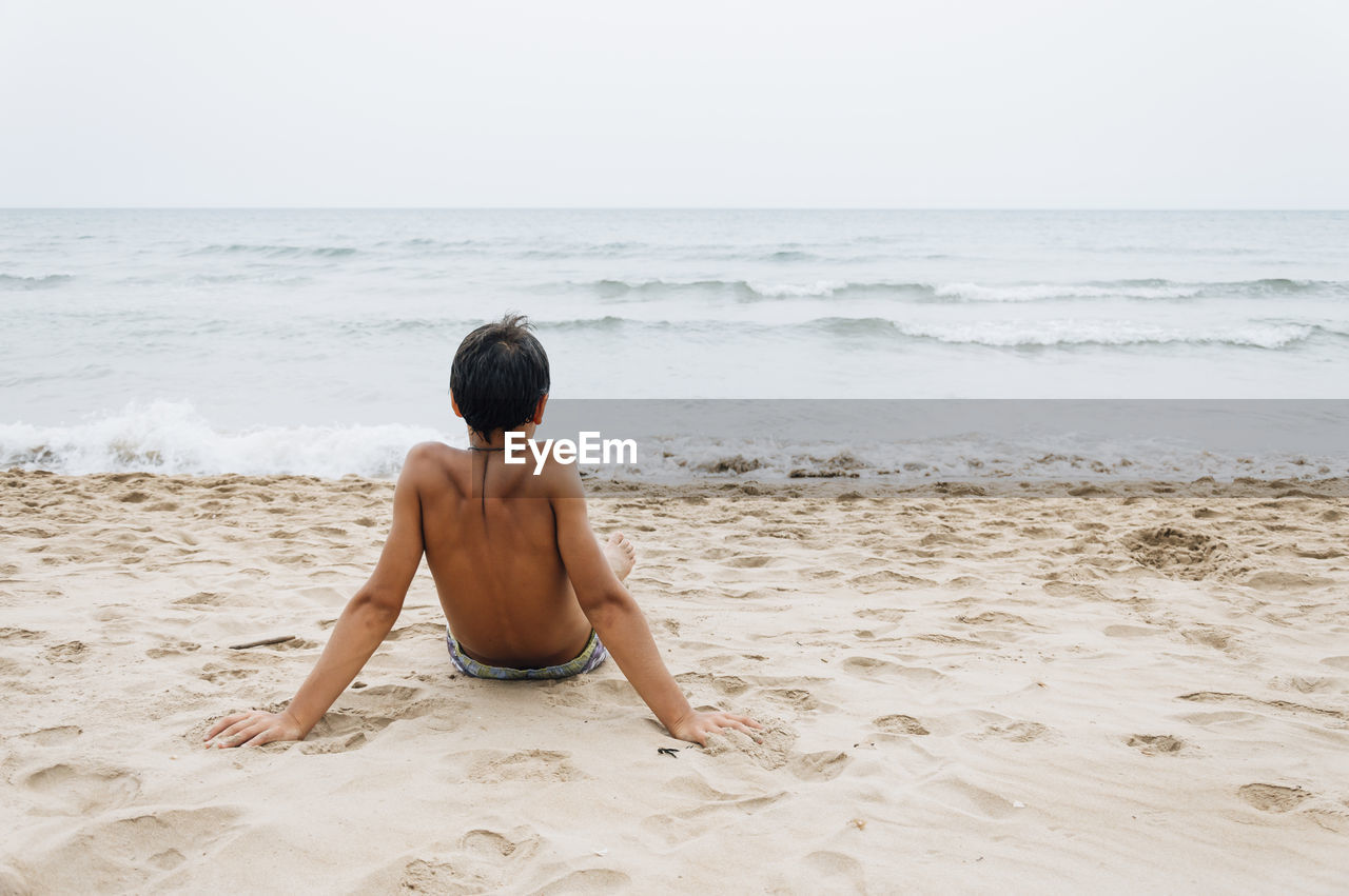 Rear view of shirtless young man relaxing at beach against clear sky