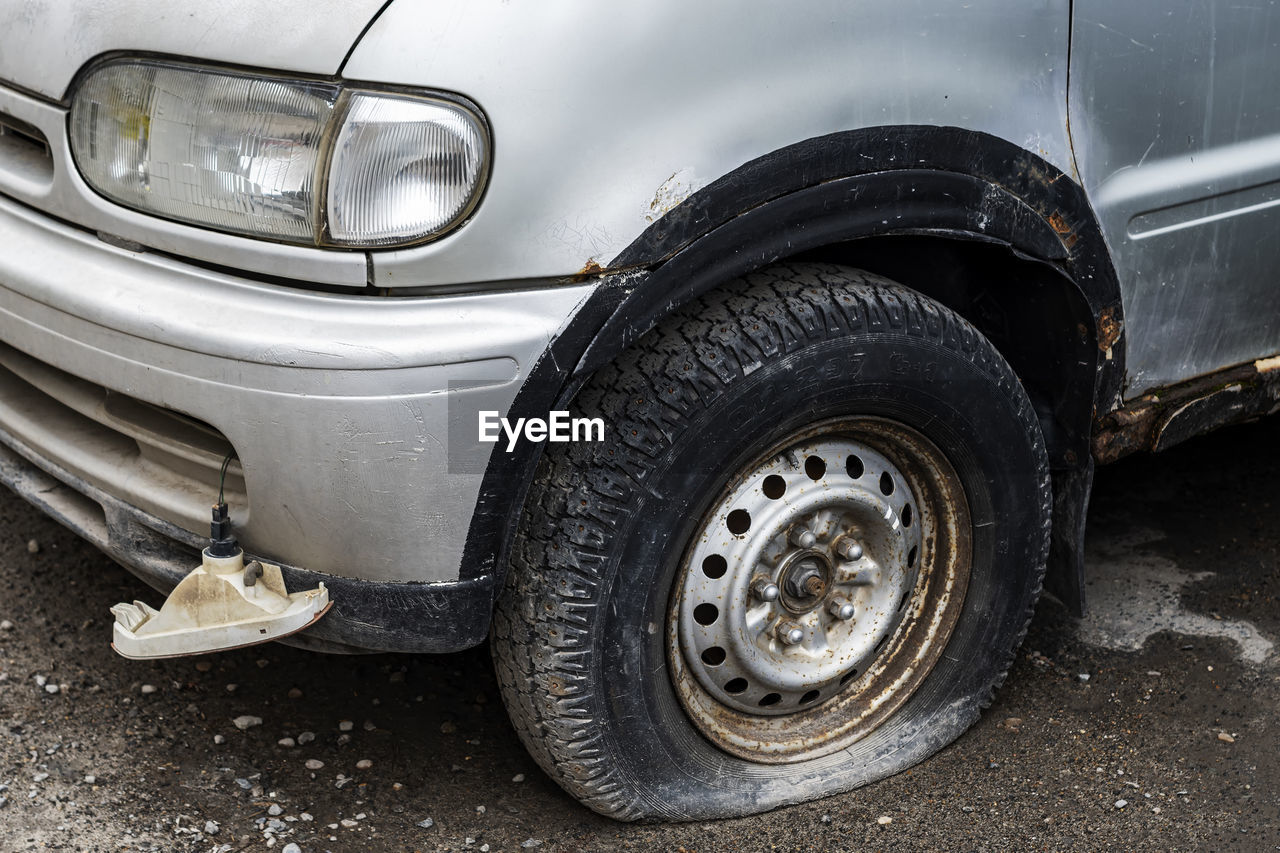 Gray car is old, with a punctured wheel and a damaged fog headlight.