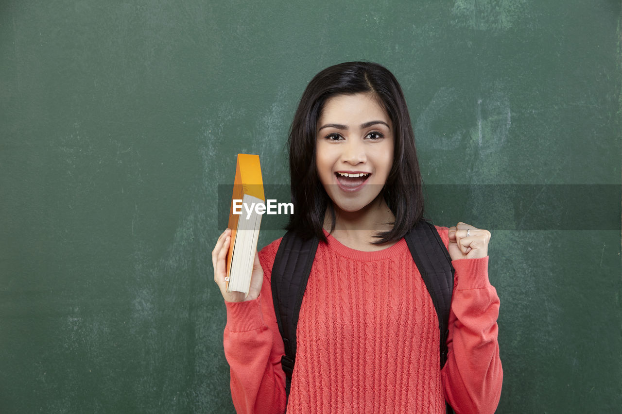 Portrait of cheerful female student gesturing while standing by blackboard