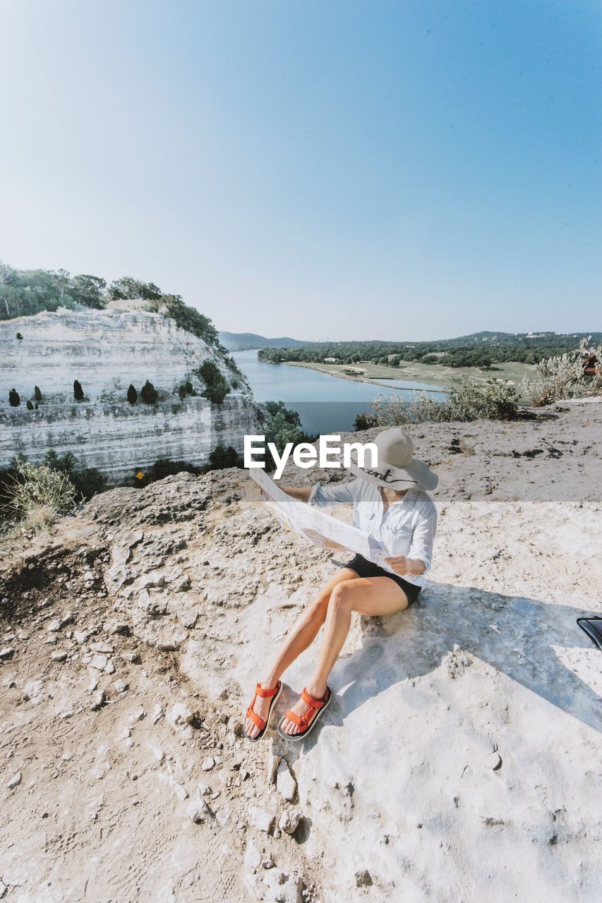 Woman reading map while sitting on rock formation against clear sky